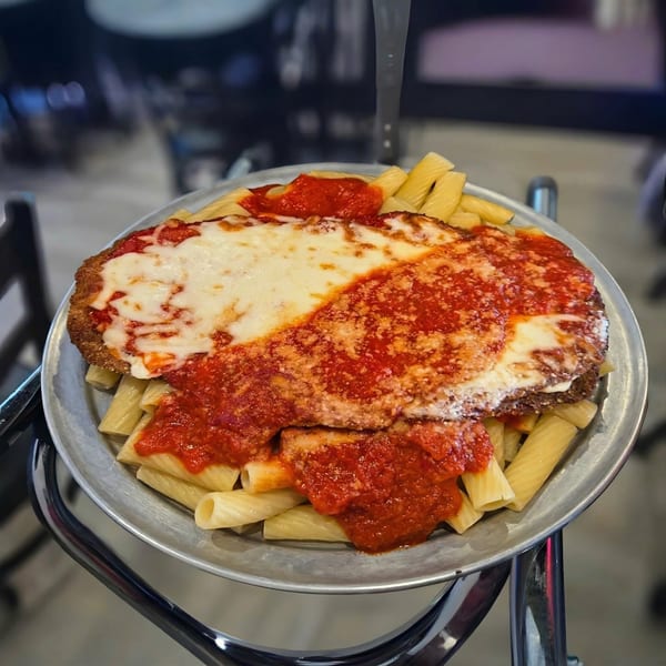 Chicken parmesan with melted cheese and red sauce, served over rigatoni pasta on a metal plate.