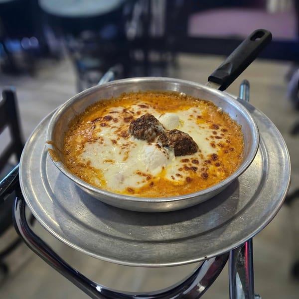 A sizzling skillet of baked meatballs and cheese in a rich tomato sauce, served on a metal stand.