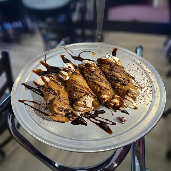 Four cannolis drizzled with chocolate sauce and powdered sugar on a round metal tray.