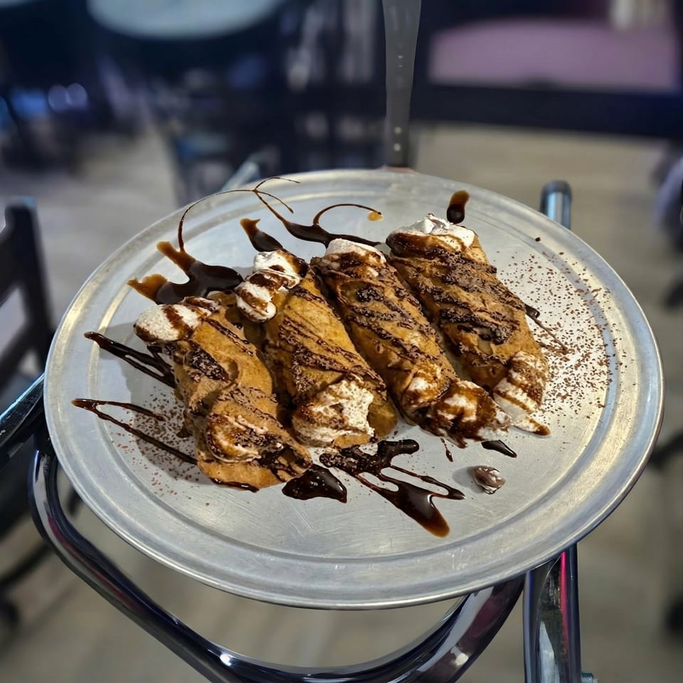 Four cannolis drizzled with chocolate sauce and powdered sugar on a round metal tray.
